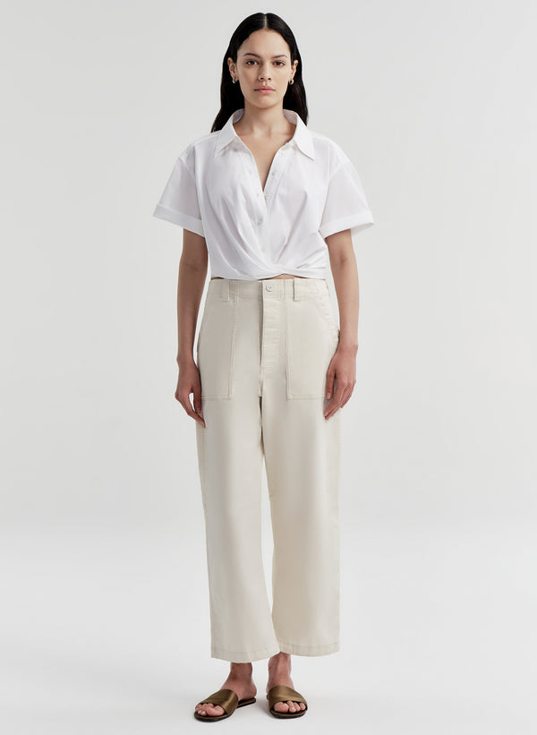 A woman stands against a light gray studio background wearing a white short-sleeve collared shirt with high-waisted cream wide-leg trousers and brown sandals.