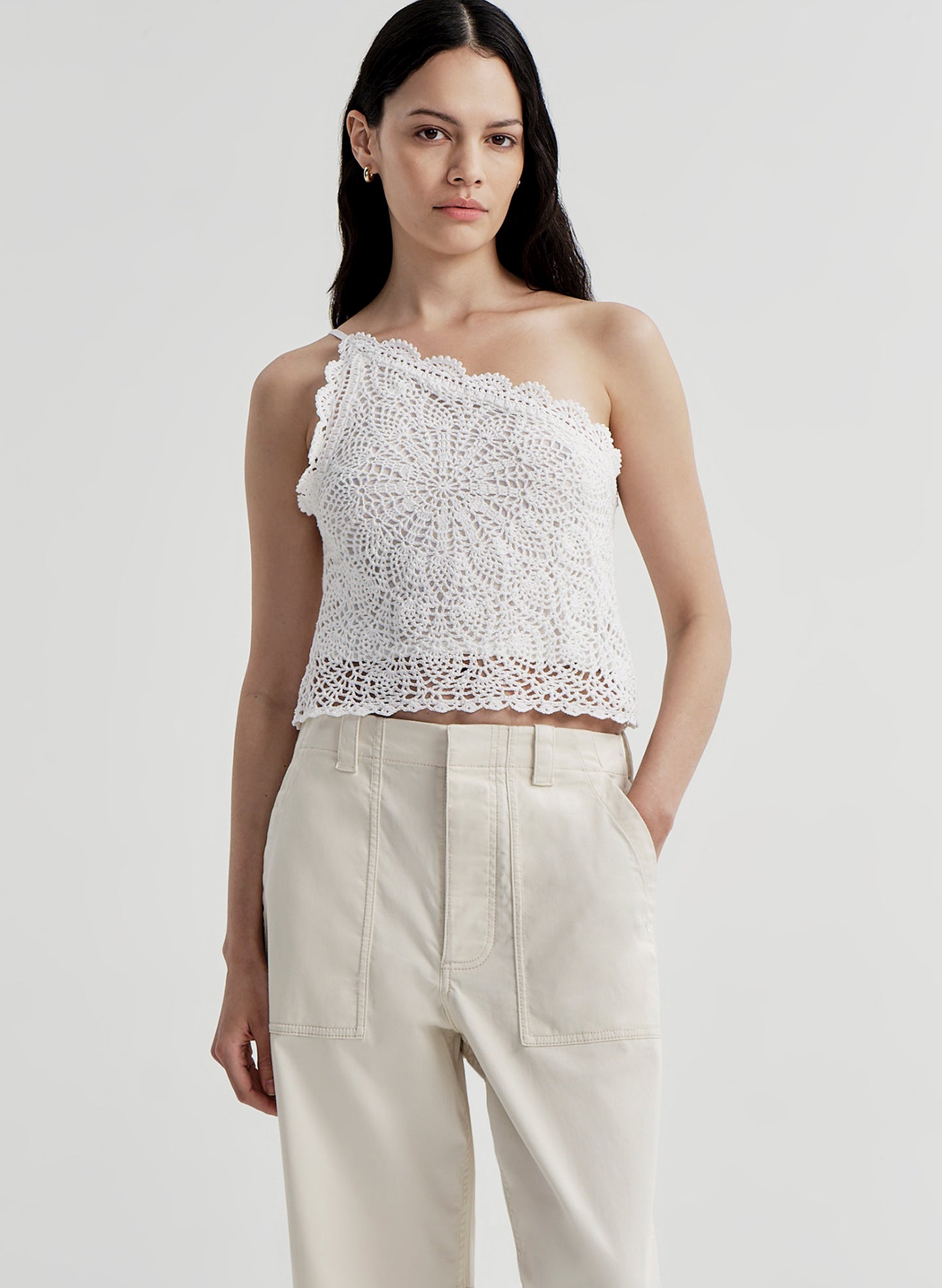 Woman wearing a white crochet one-shoulder top and beige pants on a light gray background