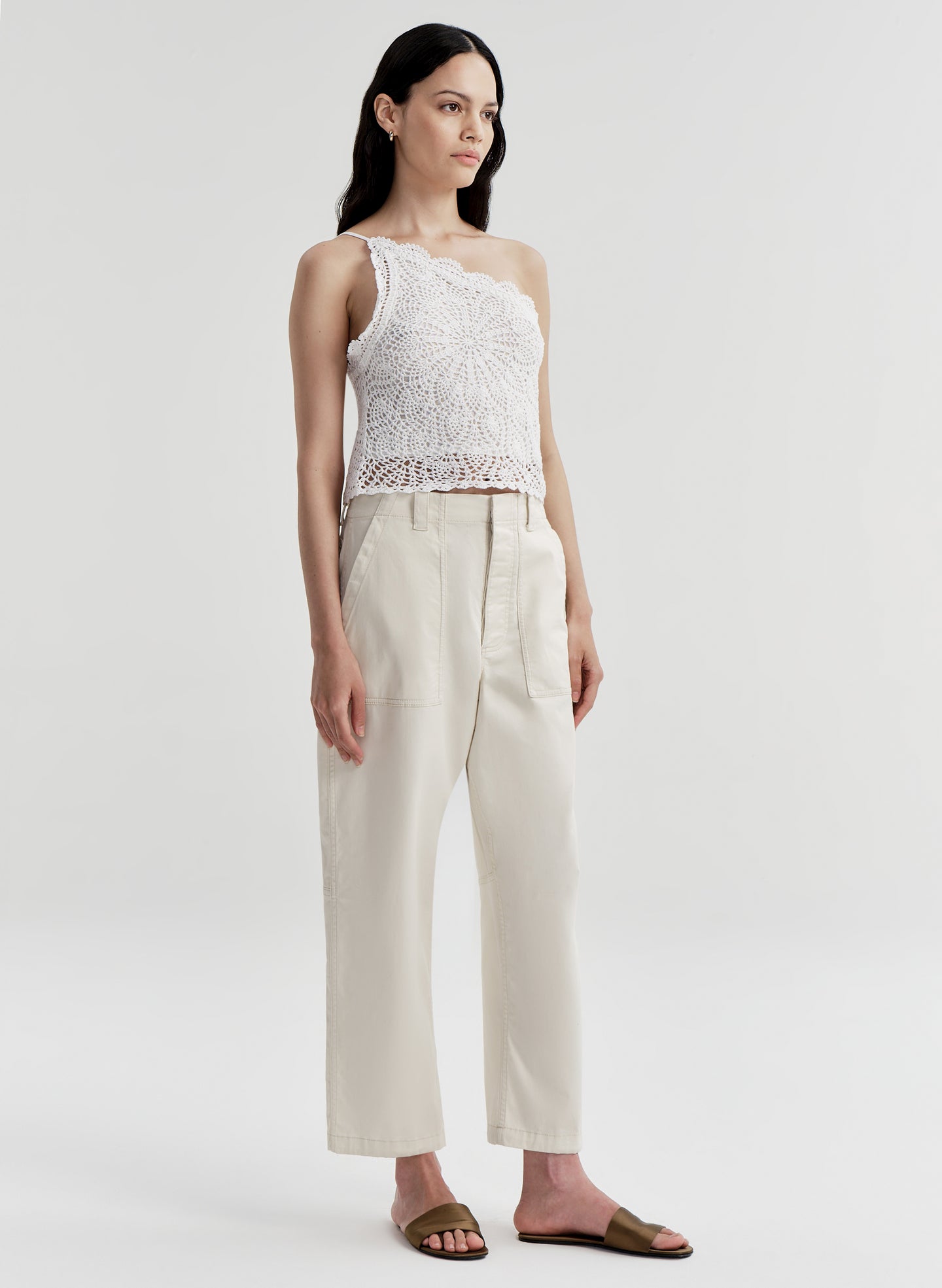 Woman wearing a white crochet one-shoulder top and beige pants on a light gray background