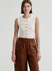 Woman wearing a white sleeveless top with buttons and brown pants on a light gray background