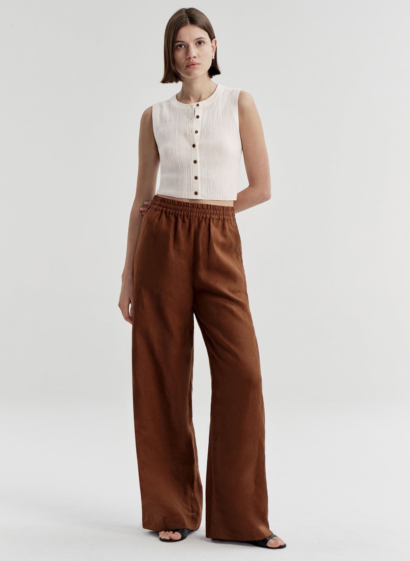 Woman wearing a white sleeveless top and brown wide-leg pants on a light gray background