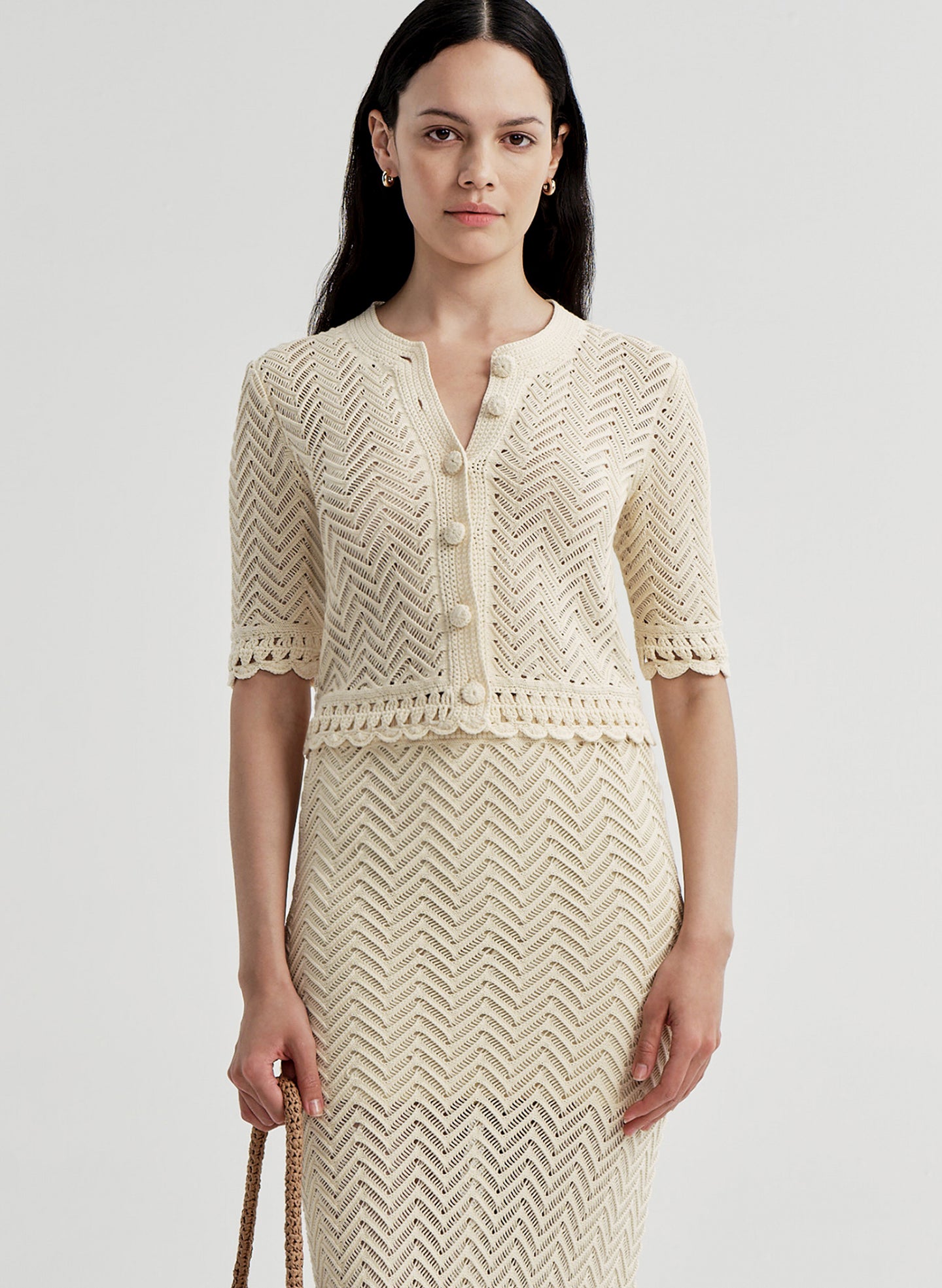 Woman wearing a beige crocheted cardigan and matching skirt on a light gray background