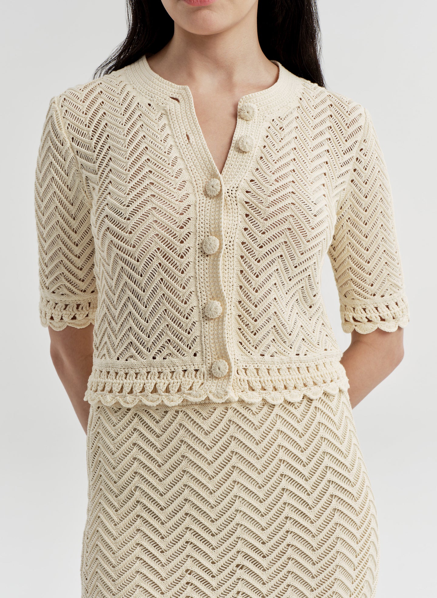 Woman wearing a beige crocheted cardigan and matching skirt on a light gray background