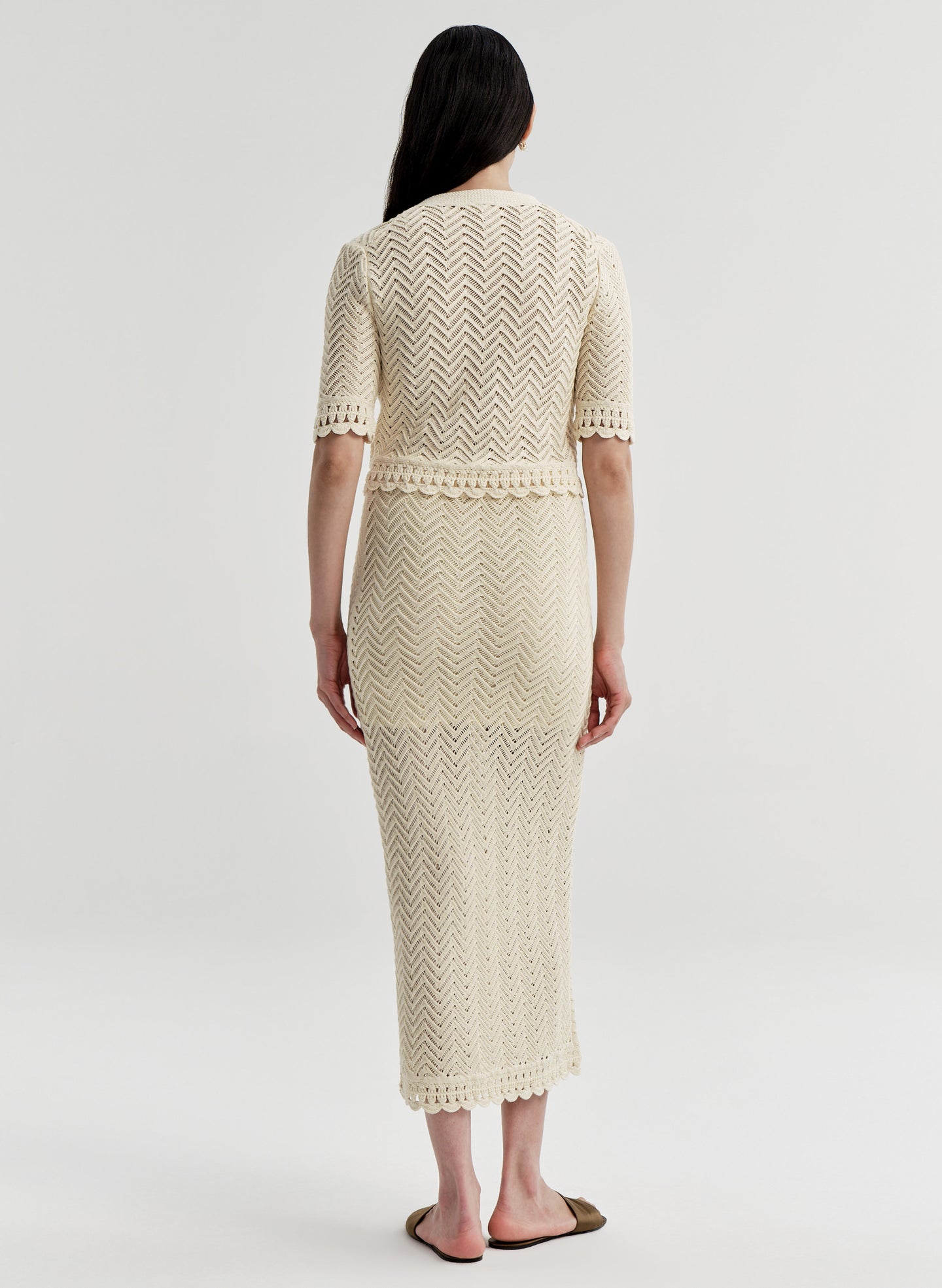 Woman wearing a beige crocheted cardigan and matching skirt on a light gray background