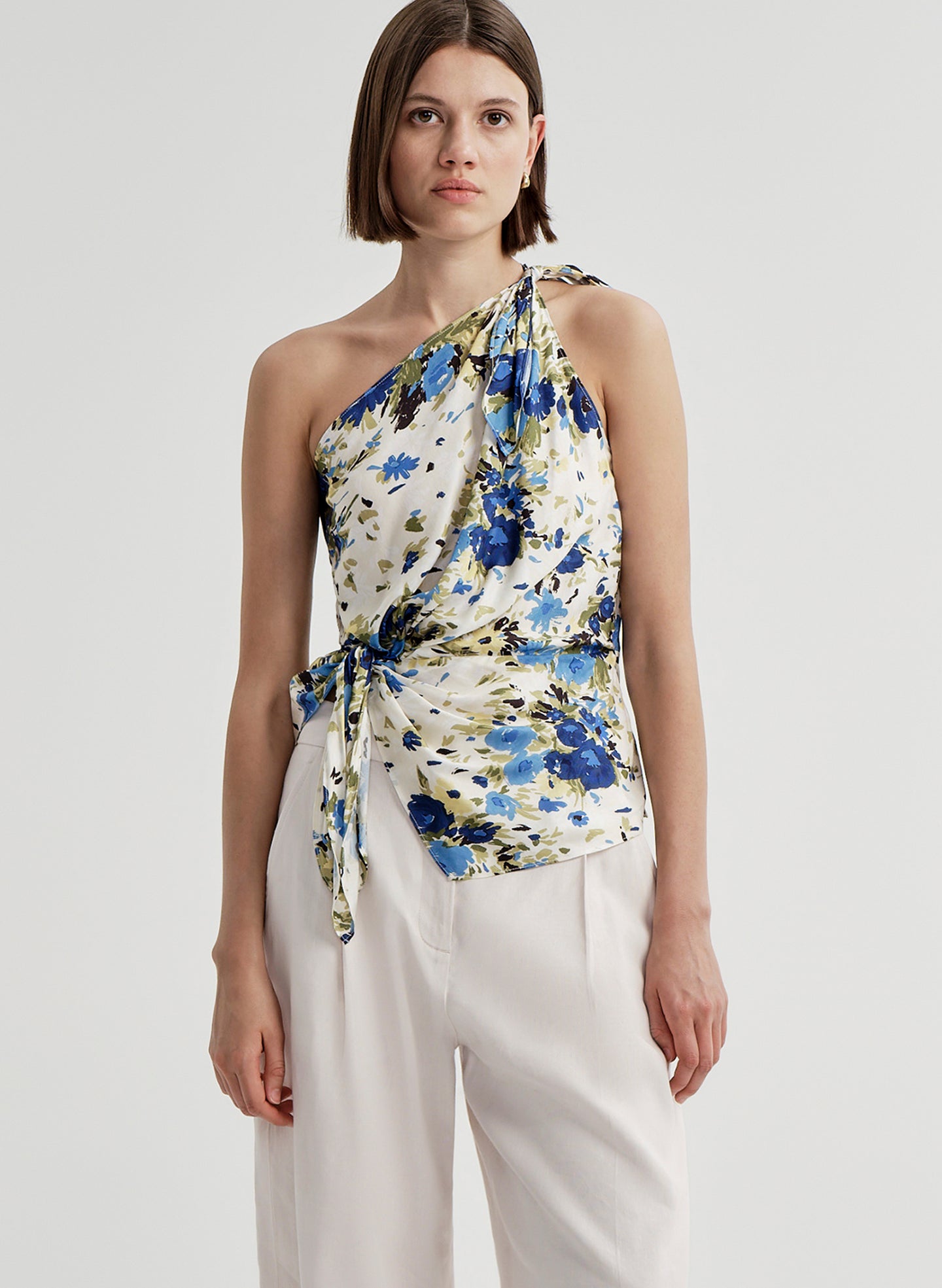 Woman wearing a floral one-shoulder top with white pants on a light gray background
