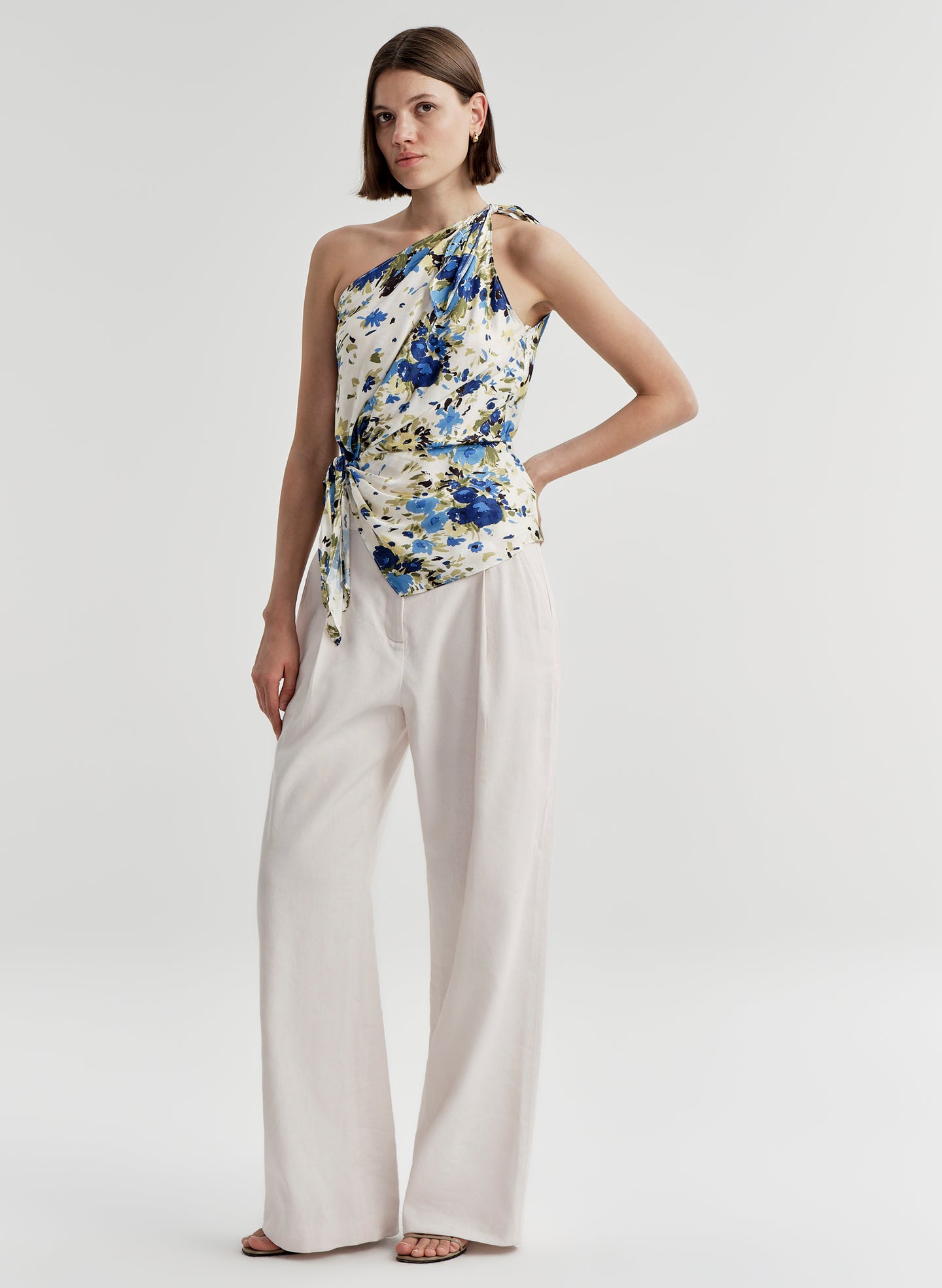 Woman wearing a floral one-shoulder top and white pants on a plain background