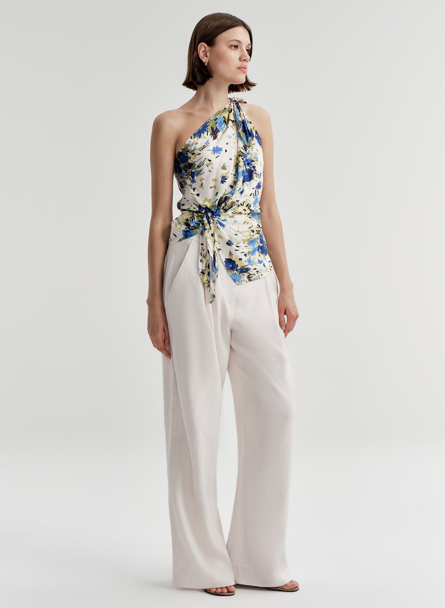 Woman wearing a one shoulder floral top and white pants on a light gray background