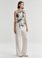 Woman wearing a one shoulder floral top and white pants on a light gray background