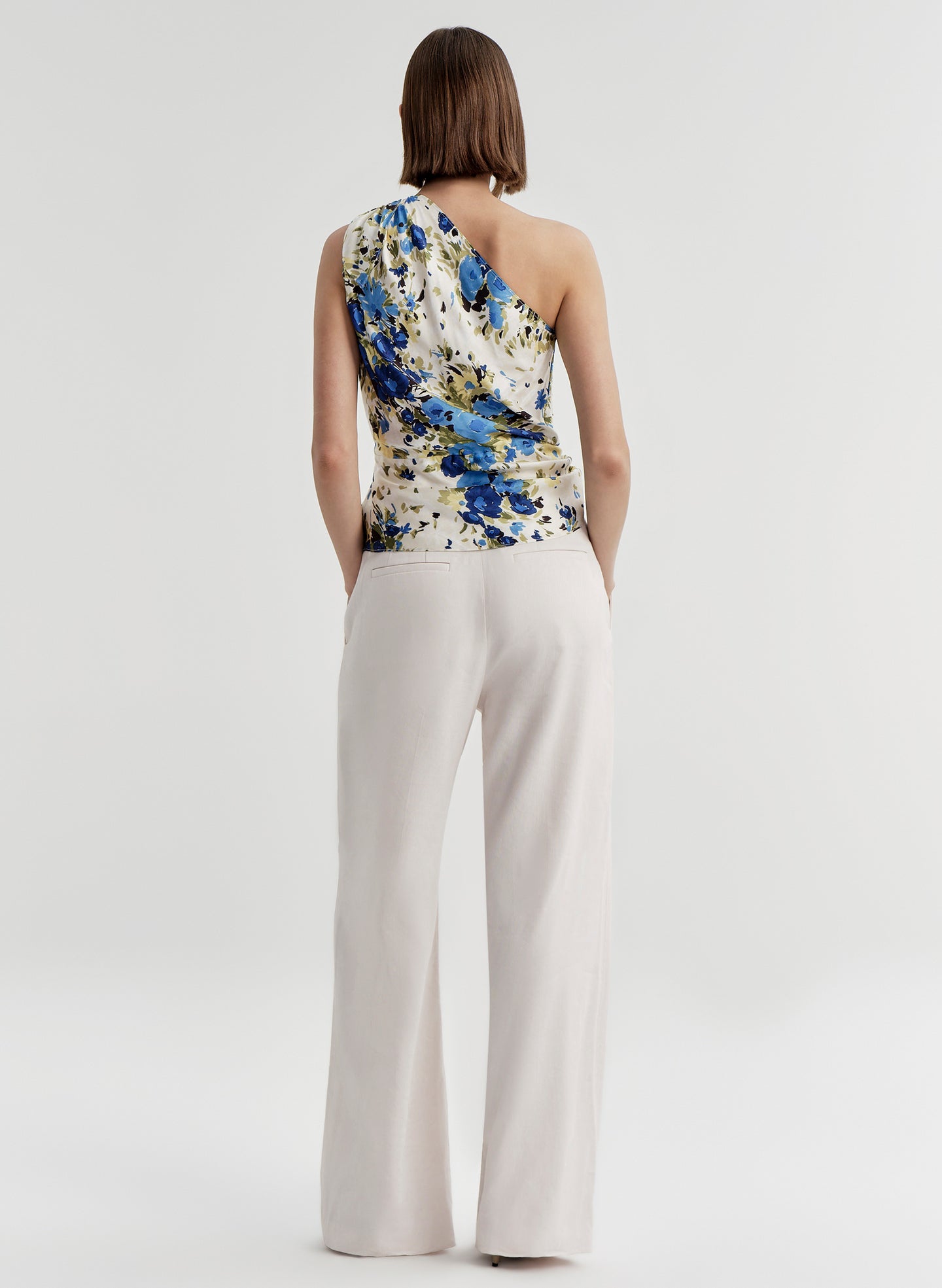 Woman wearing a floral one-shoulder top and white pants on a light gray background