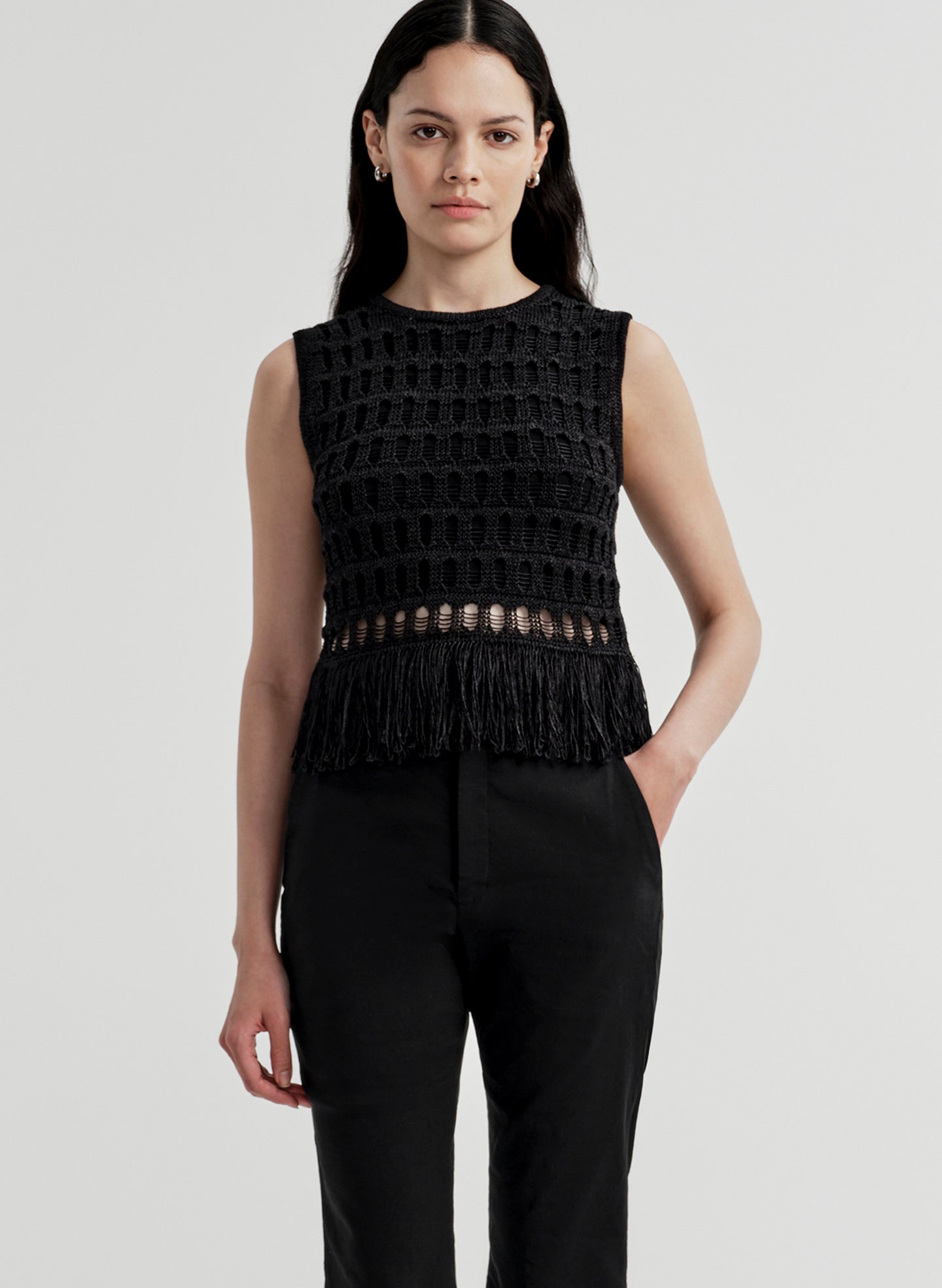 Woman wearing a black sleeveless crochet top with fringe details on a light gray background