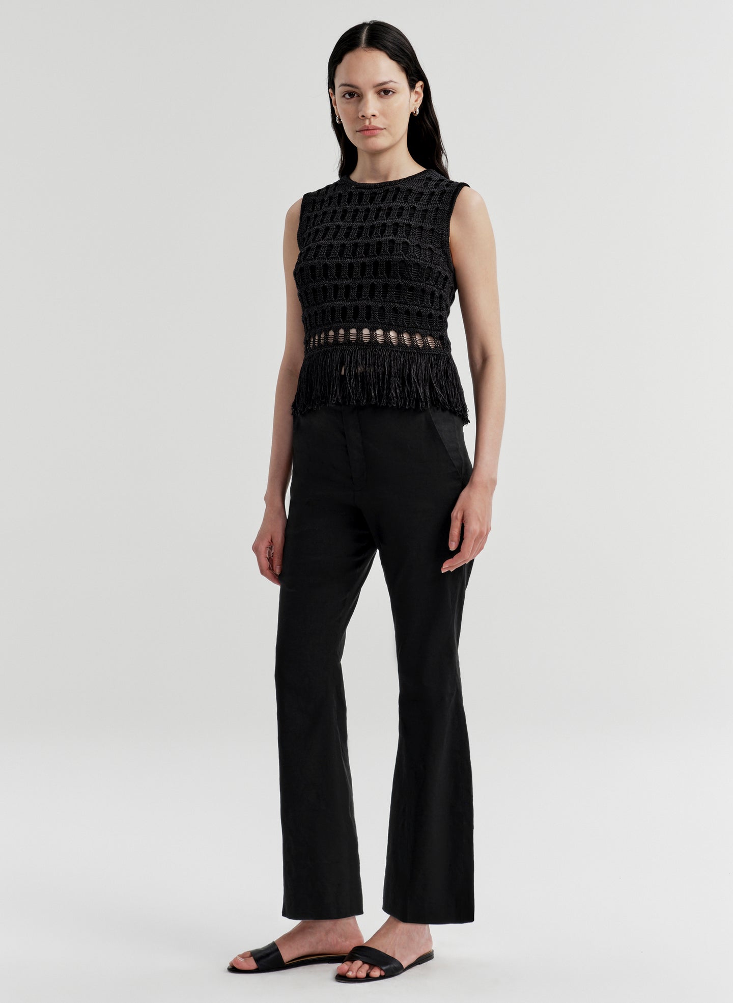 Woman wearing a black sleeveless crochet top with fringe details and black pants on a light gray background
