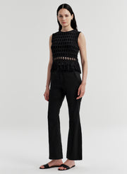 Woman wearing a black sleeveless crochet top with fringe details and black pants on a light gray background