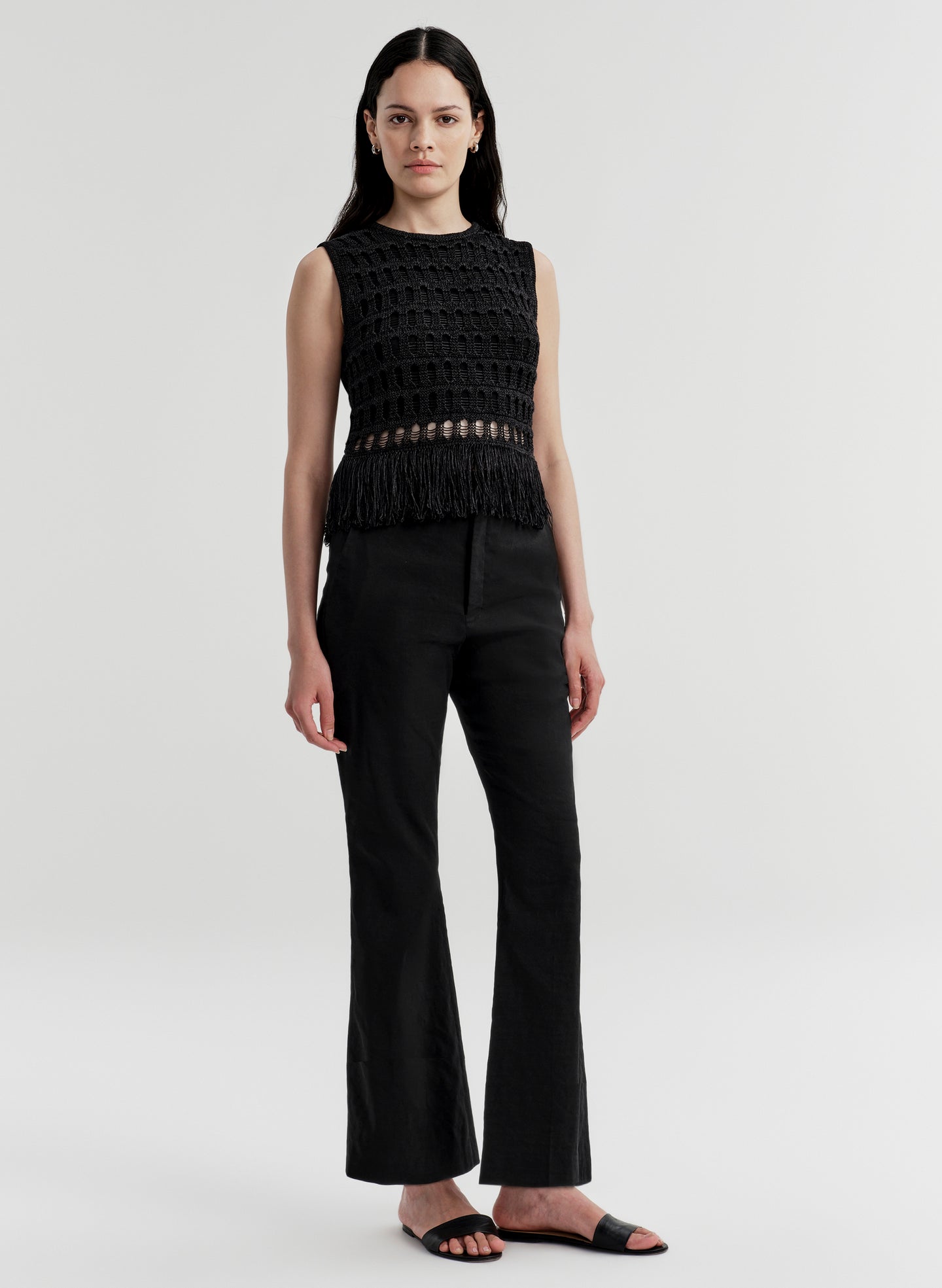 Woman wearing a black sleeveless crochet top with fringe details and black pants on a light gray background