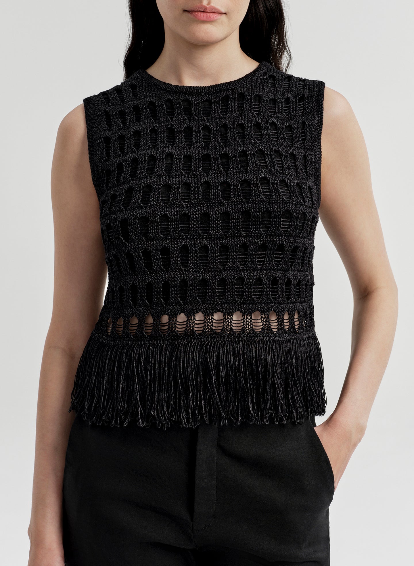 Woman wearing a black sleeveless crochet top with fringe details on a light gray background