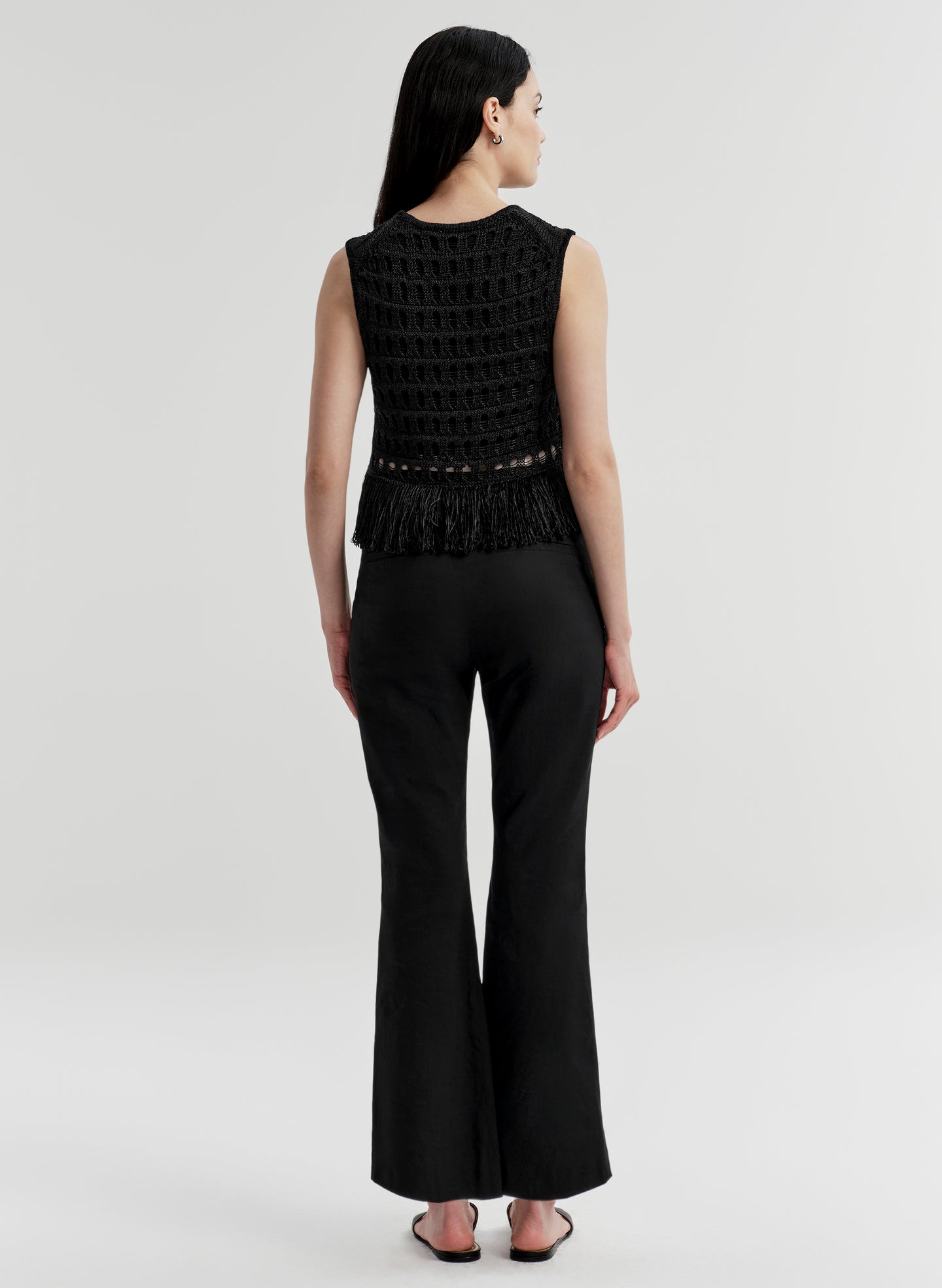 Woman wearing a black sleeveless crochet top with fringe details and black pants on a light gray background