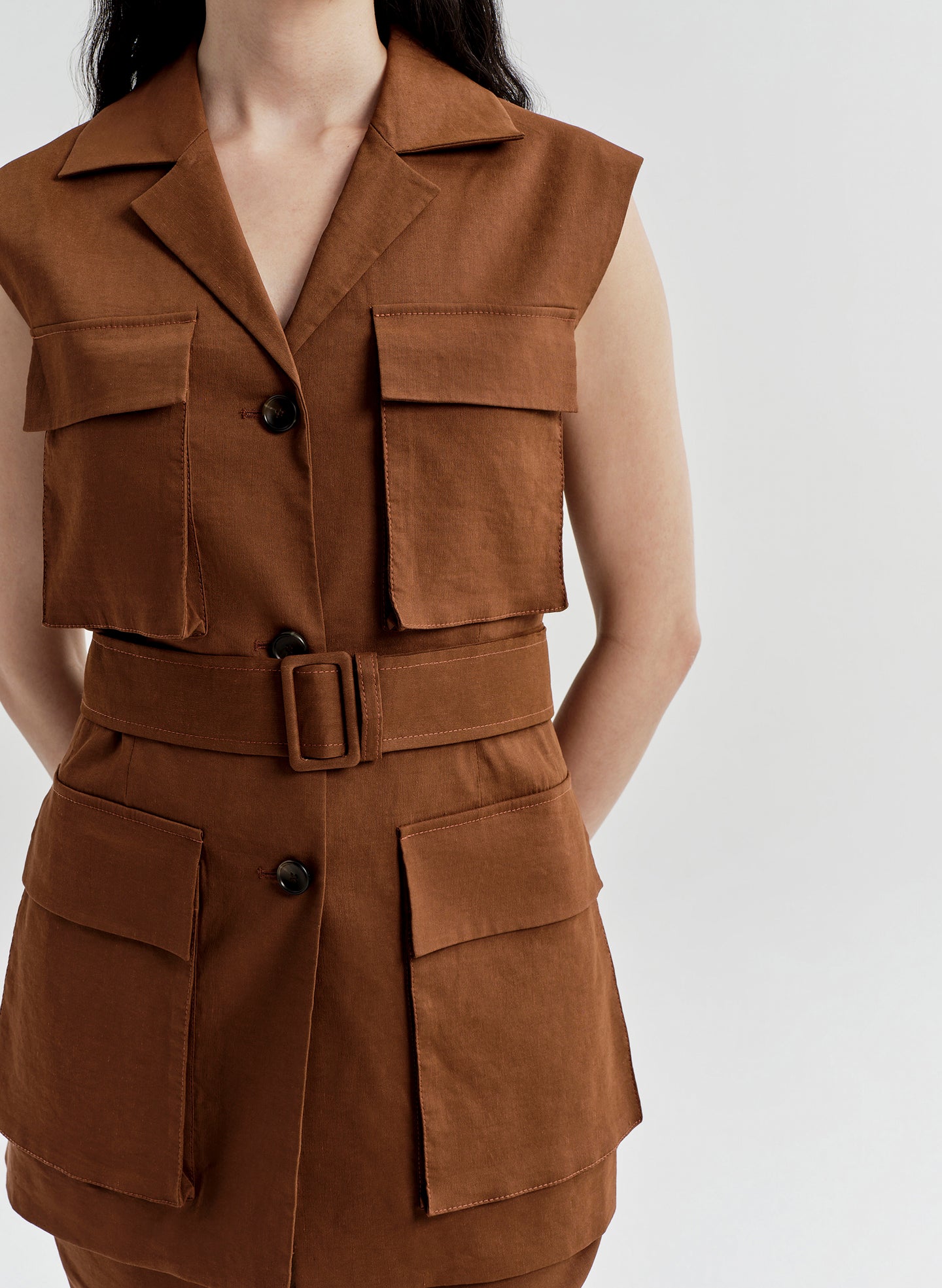 Woman wearing a brown sleeveless vest jacket with a belt and pockets on a white background