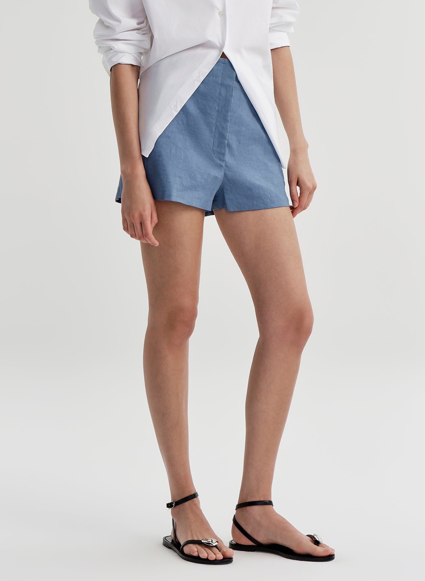 Woman wearing blue linen shorts and a white button up shirt on a light gray background