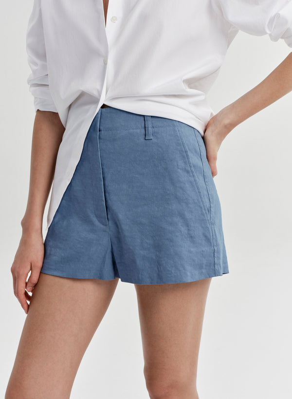 Woman wearing blue linen shorts and a white button up shirt on a light gray background