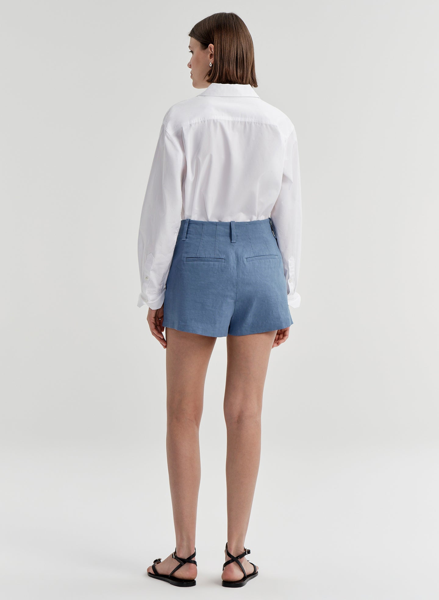 Woman wearing blue linen shorts and a white tucked in shirt on a light gray background