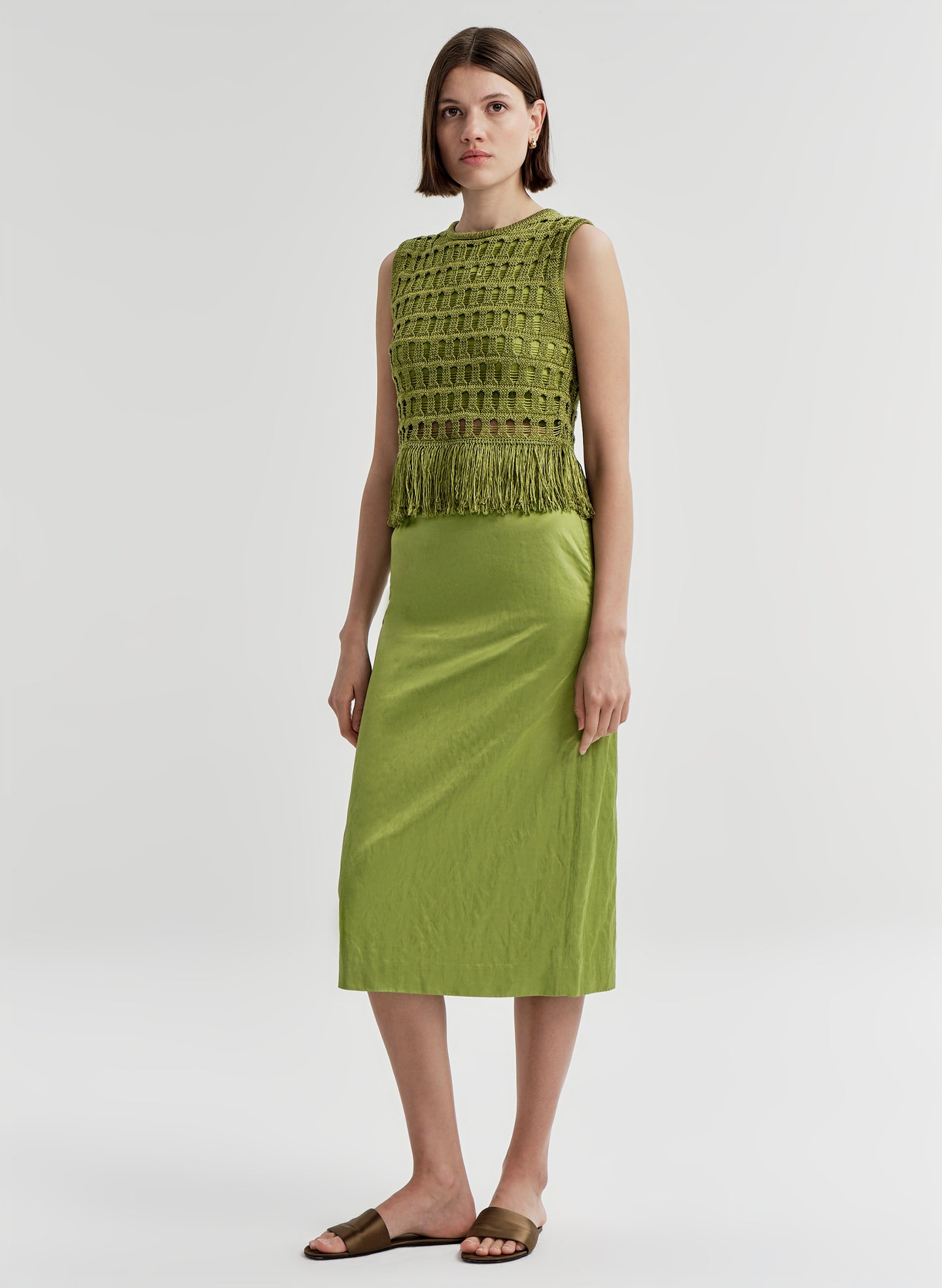 Woman wearing a crochet green sleeveless top with fringe details with a green skirt on a light gray background