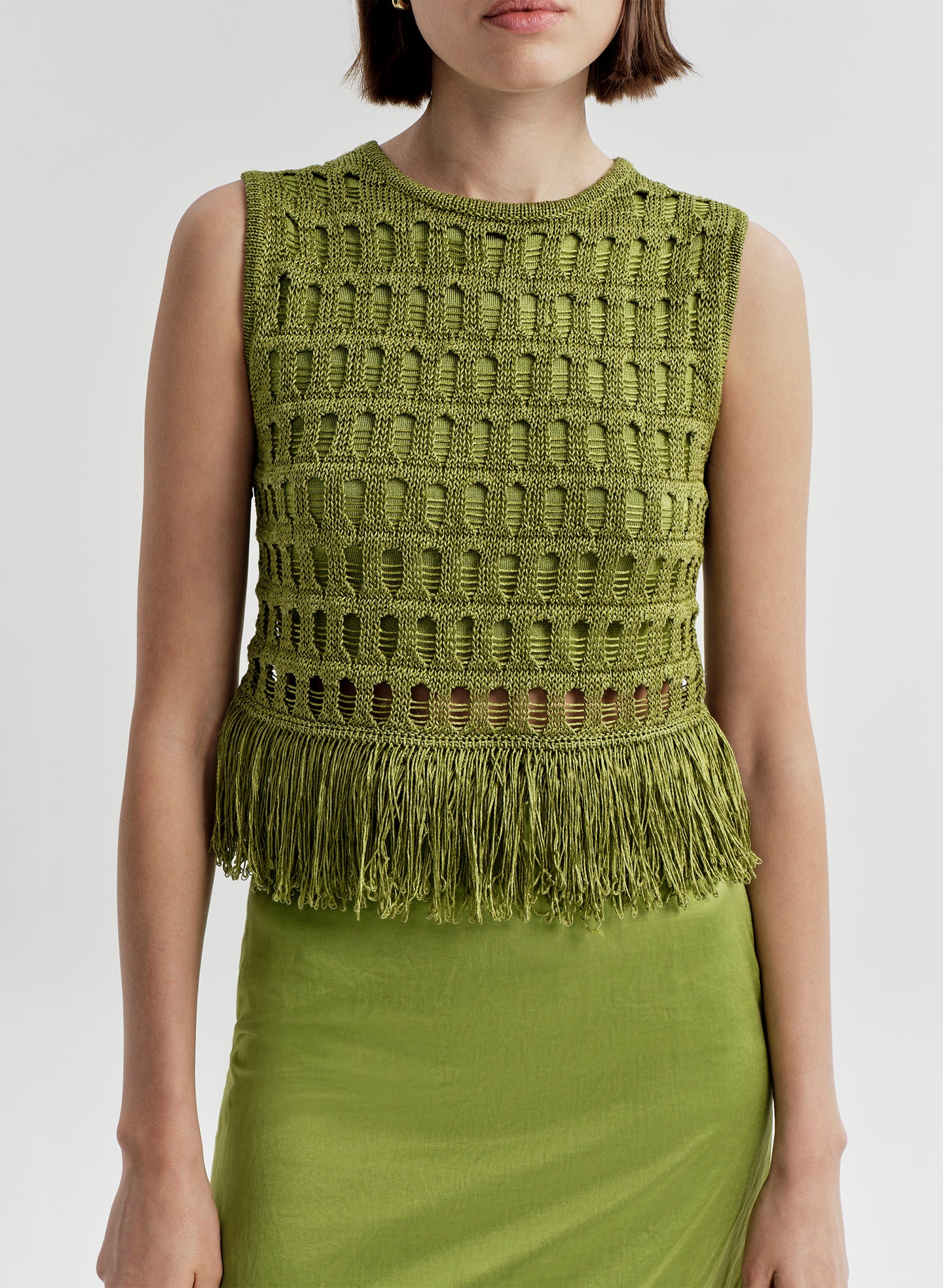 Woman wearing a crochet green sleeveless top with fringe details on a light gray background