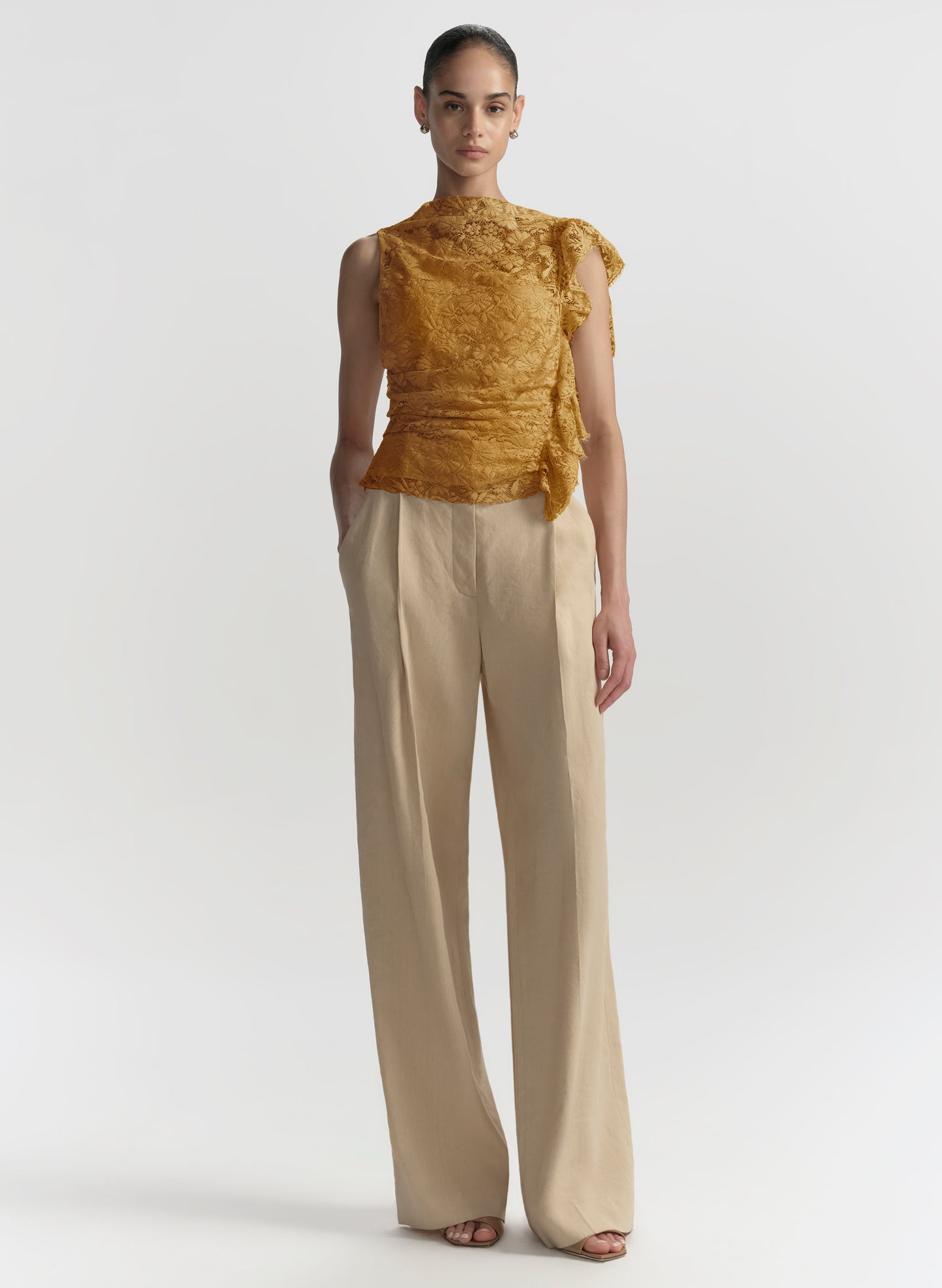 A model stands against a light gray studio background wearing a sleeveless textured mustard yellow top with ruffled shoulder details and high-necked, wide-leg beige trousers.