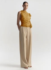 A model stands against a light gray studio background wearing a sleeveless textured mustard-yellow top with ruffle details and high-waisted, wide-leg beige trousers.