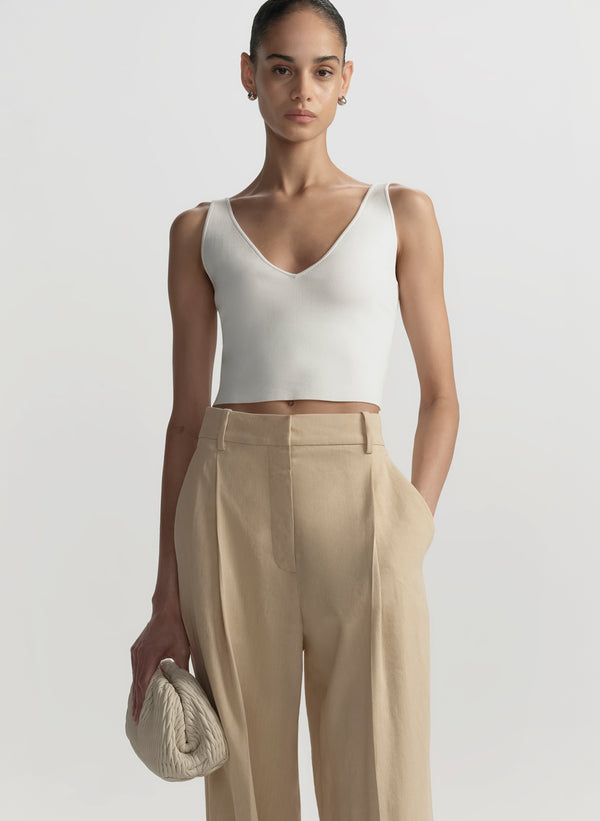 Woman wearing a white sleeveless top and beige pants against a plain background