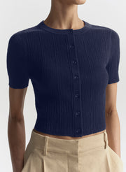 A woman wearing a navy blue short-sleeve ribbed knit cardigan top with a button front, paired with beige trousers, shown against a light gray studio background.