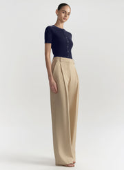 A model stands in a studio wearing a dark navy short-sleeve knit top with a button placket and high-waisted beige wide-leg trousers against a light gray background.