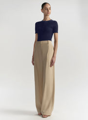 A model stands against a light gray studio background wearing a short-sleeve navy knit top and high-waisted, wide-leg beige trousers.