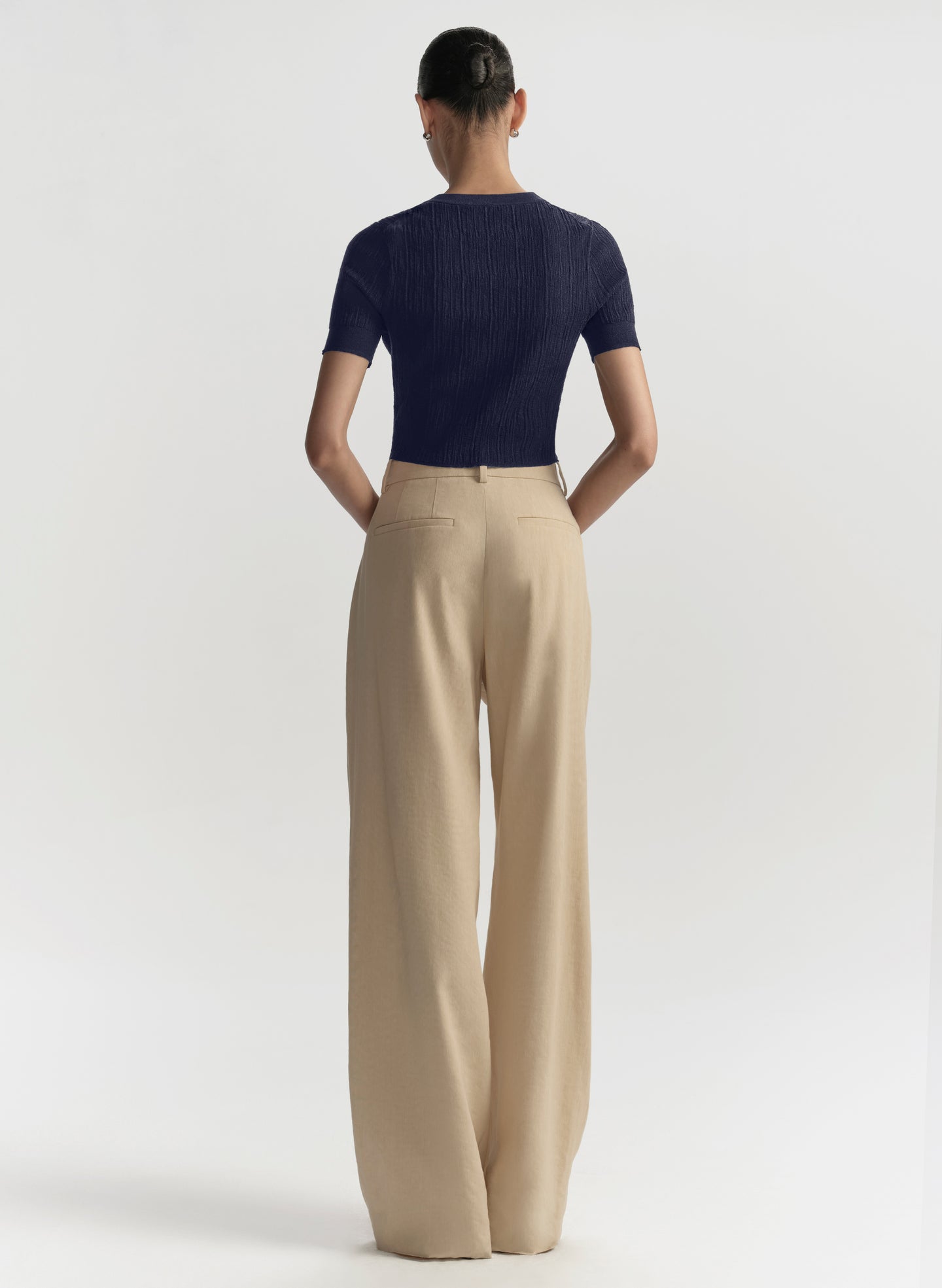 A model shown from the back wearing a navy blue short-sleeve ribbed knit top and beige wide-leg trousers, photographed against a clean white studio background.