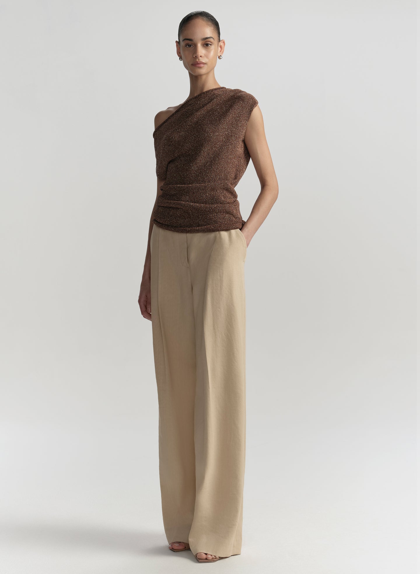 Woman wearing a brown sleeveless top and beige pants on a light gray background