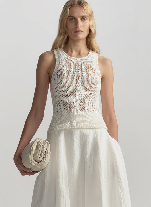 Woman wearing a white sleeveless knitted top and holding a white bag against a plain background
