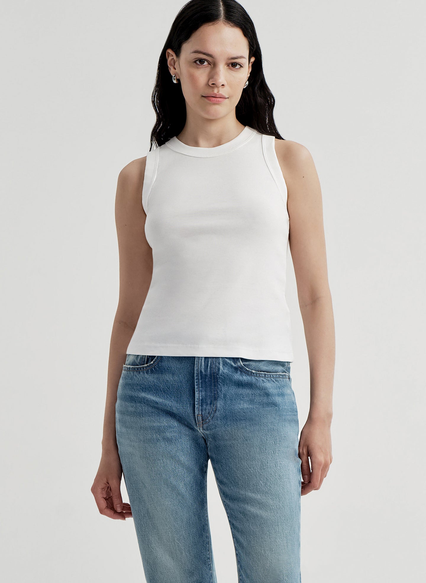 Woman wearing a white sleeveless top and blue jeans on a light gray background