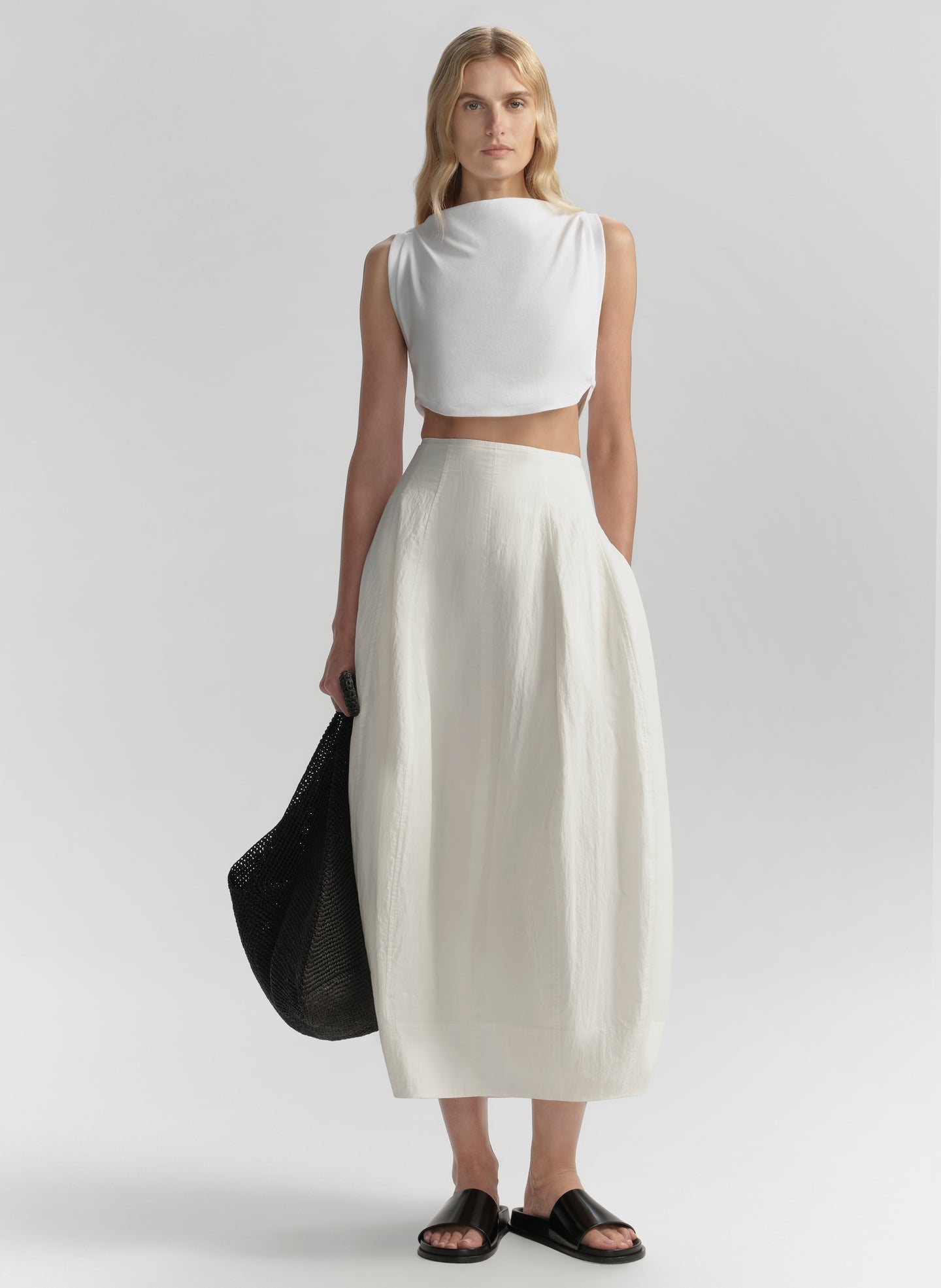 Woman wearing a white sleeveless top and white skirt holding a black bag on a light gray background