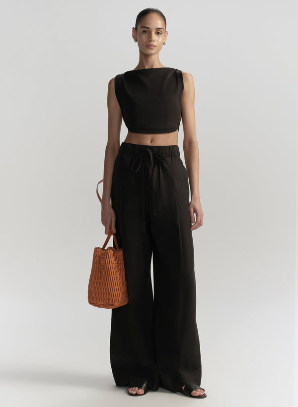 Woman wearing a black sleeveless top and wide-leg pants, holding an orange handbag on a white background.