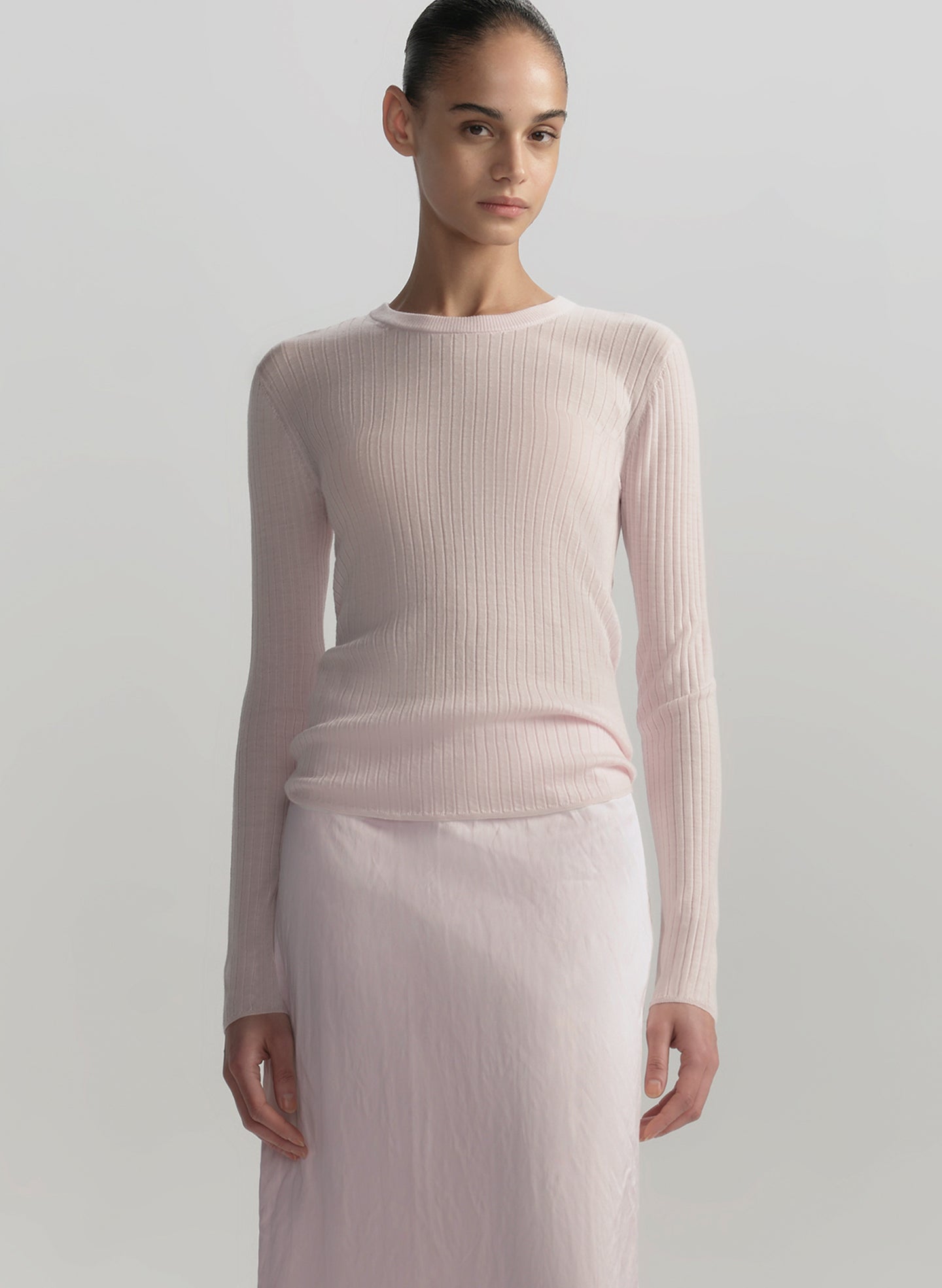 Woman wearing a light pink ribbed sweater and white pants against a plain background