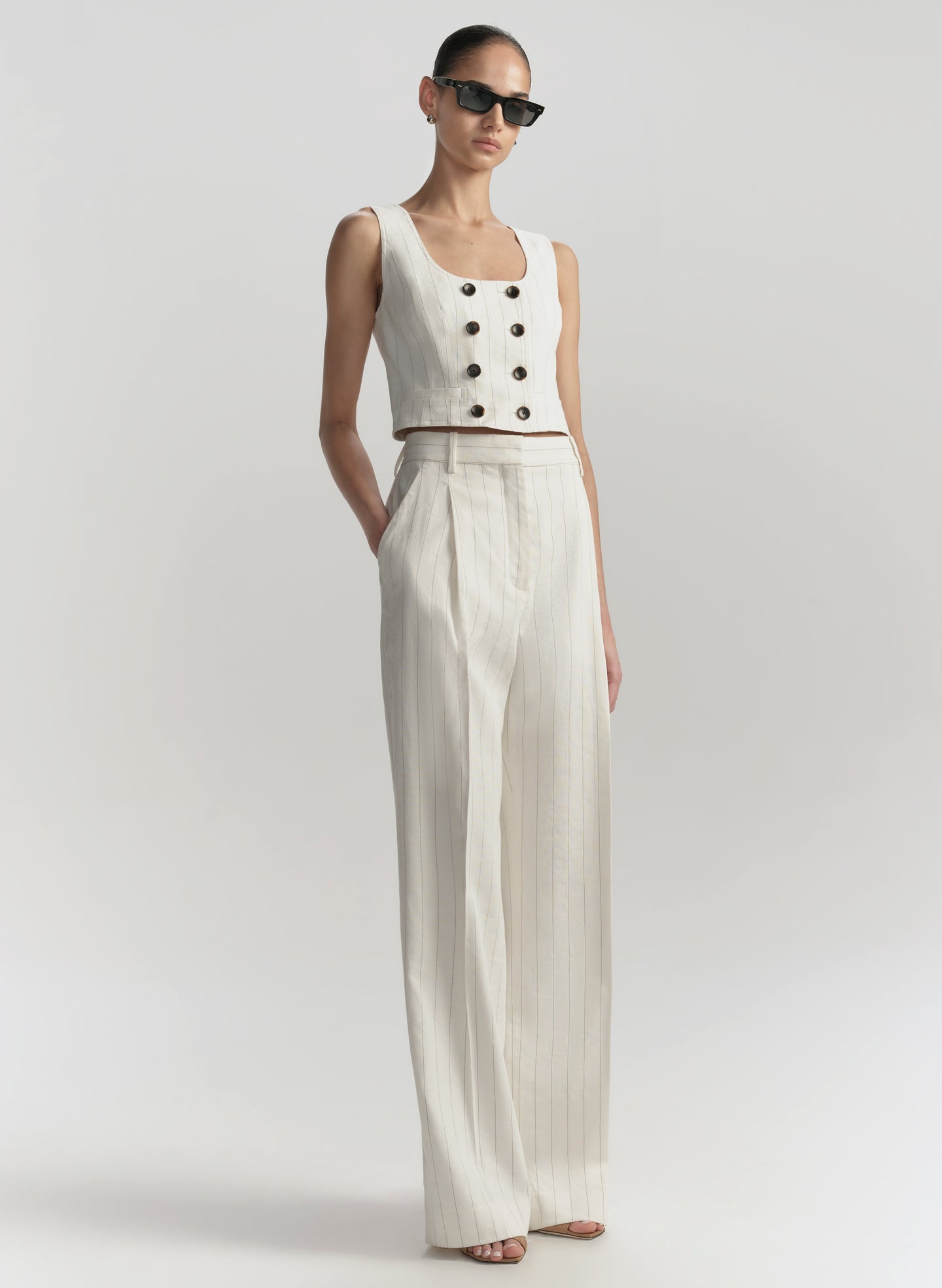 Woman wearing a white sleeveless top with buttons and wide-leg pants on a light gray background
