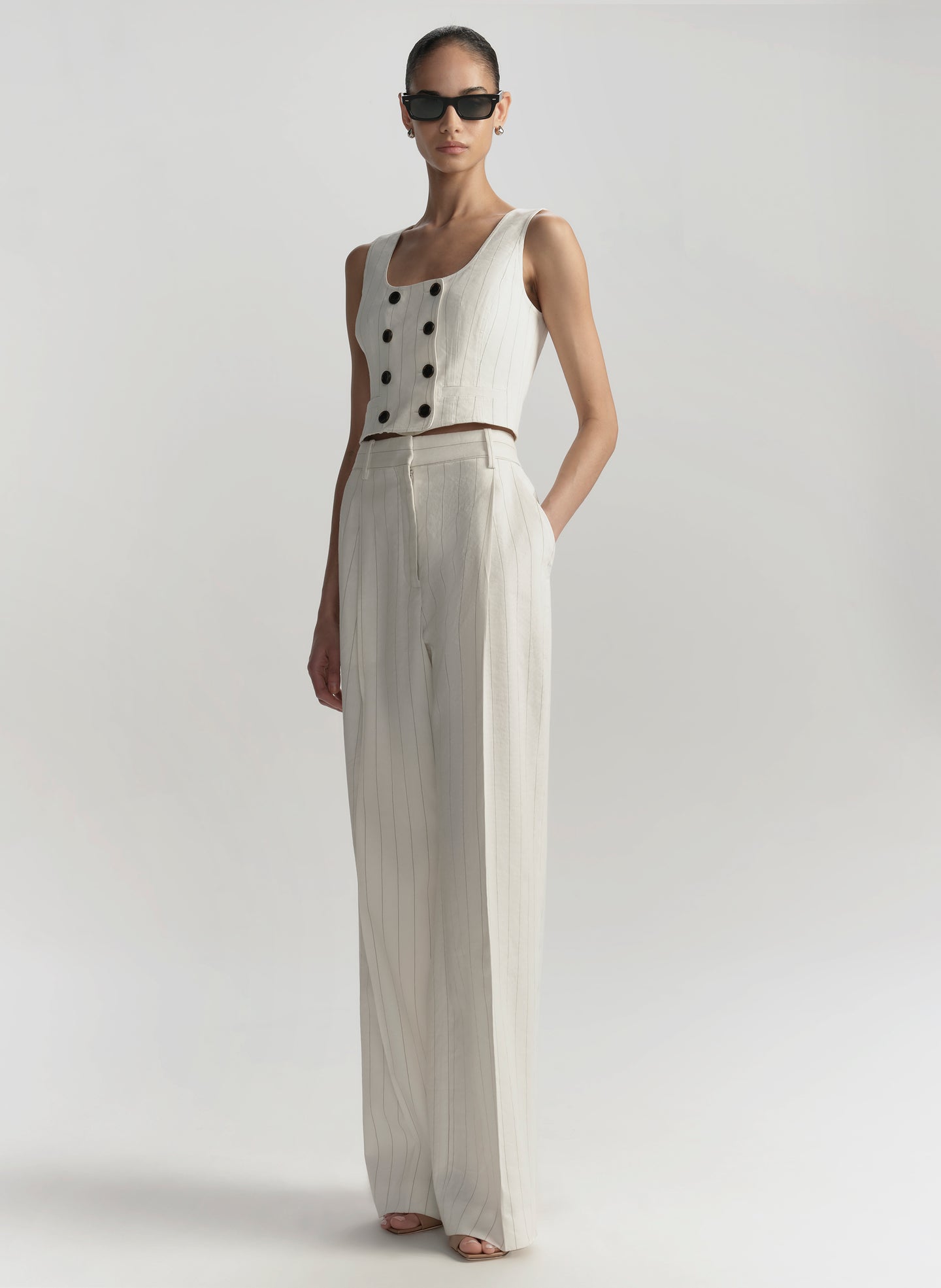Woman wearing a white sleeveless top with buttons and wide-leg pants on a plain background