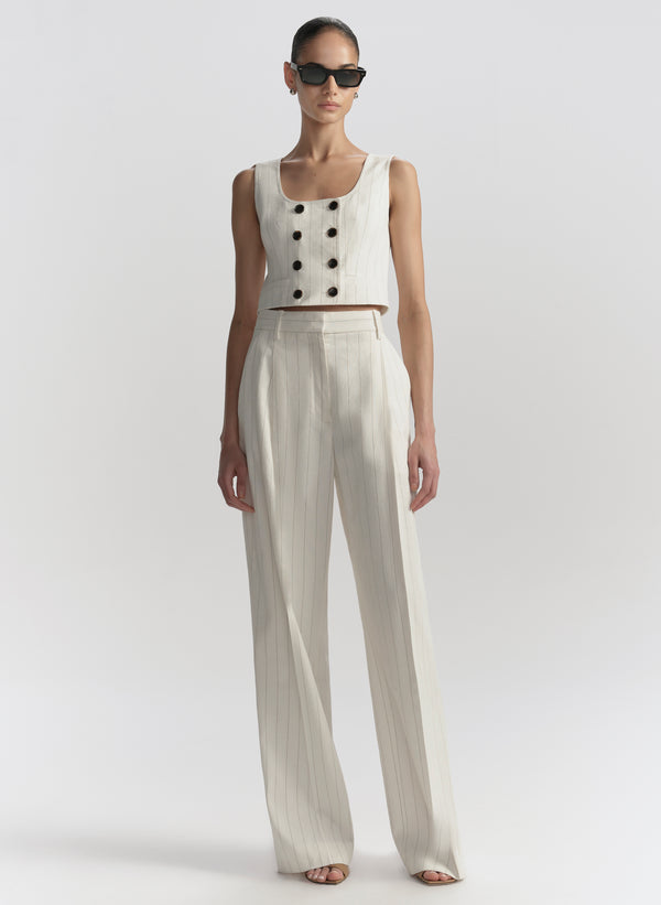 Woman wearing a white sleeveless top with black buttons and beige wide-leg pants on a light gray background