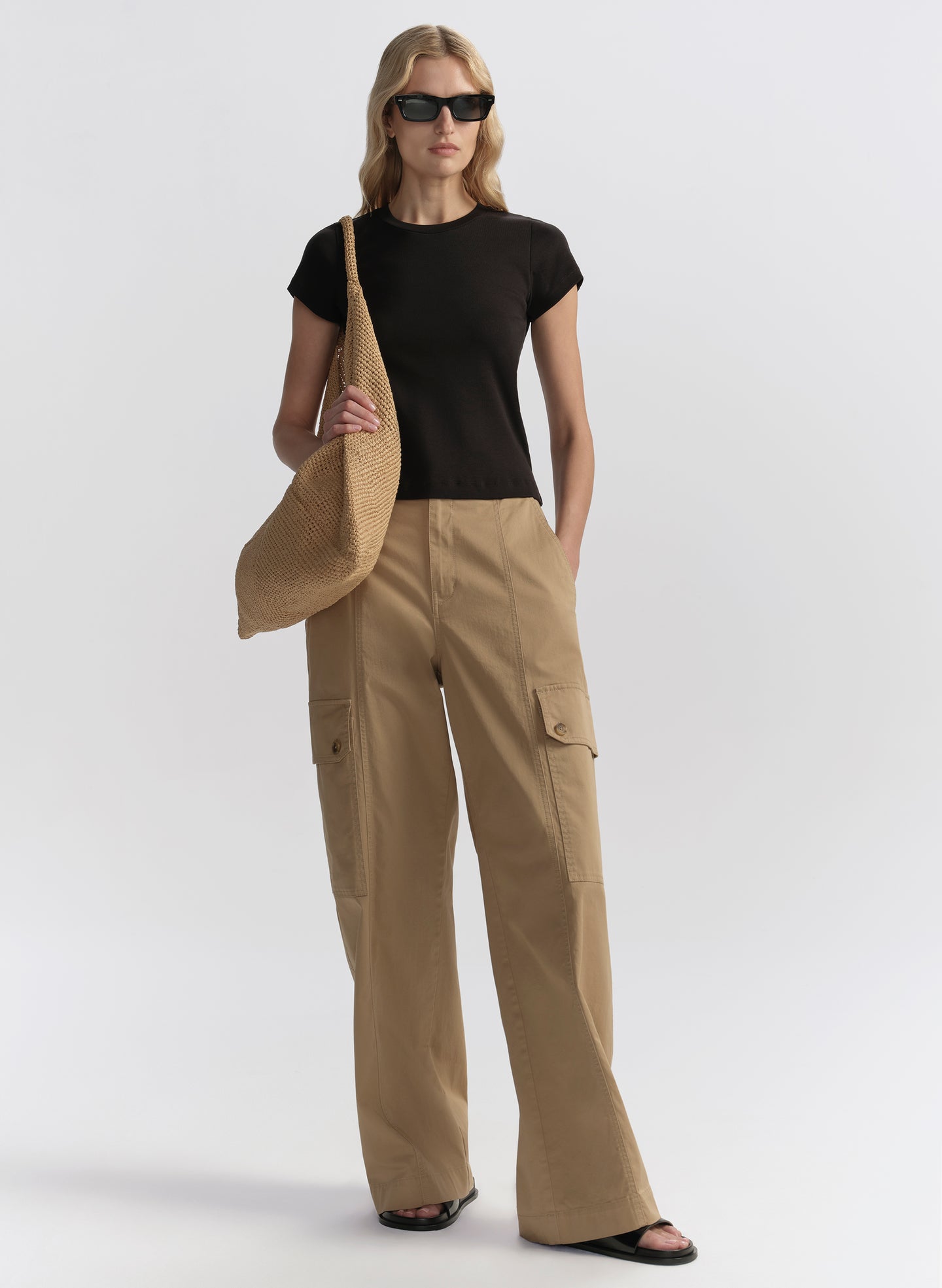 Woman wearing beige cargo pants and a black top, holding a beige bag on a white background