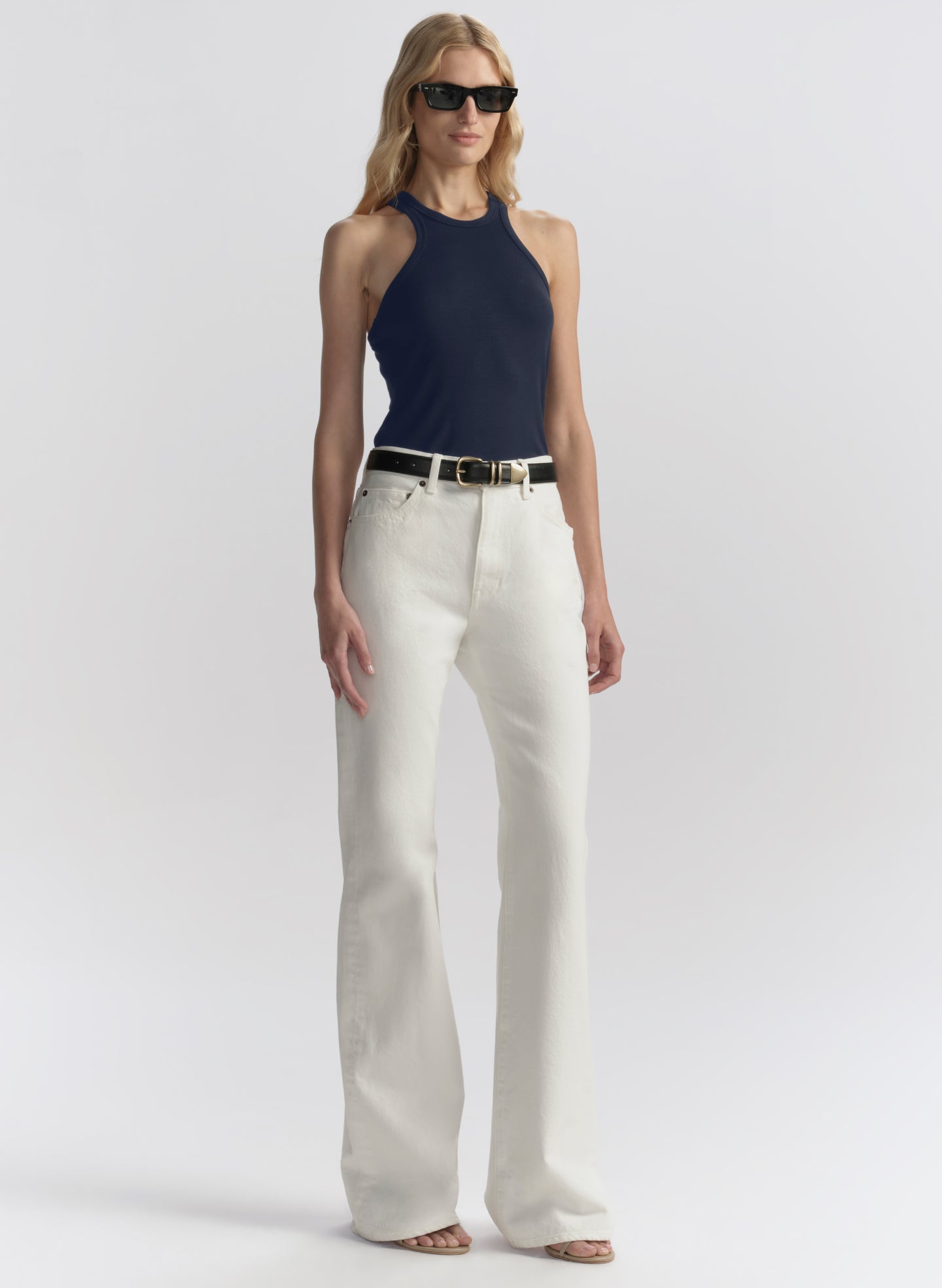 A model wearing a navy blue tank top, off-white wide-leg jeans with a black belt, and black sunglasses, standing against a light gray studio background.