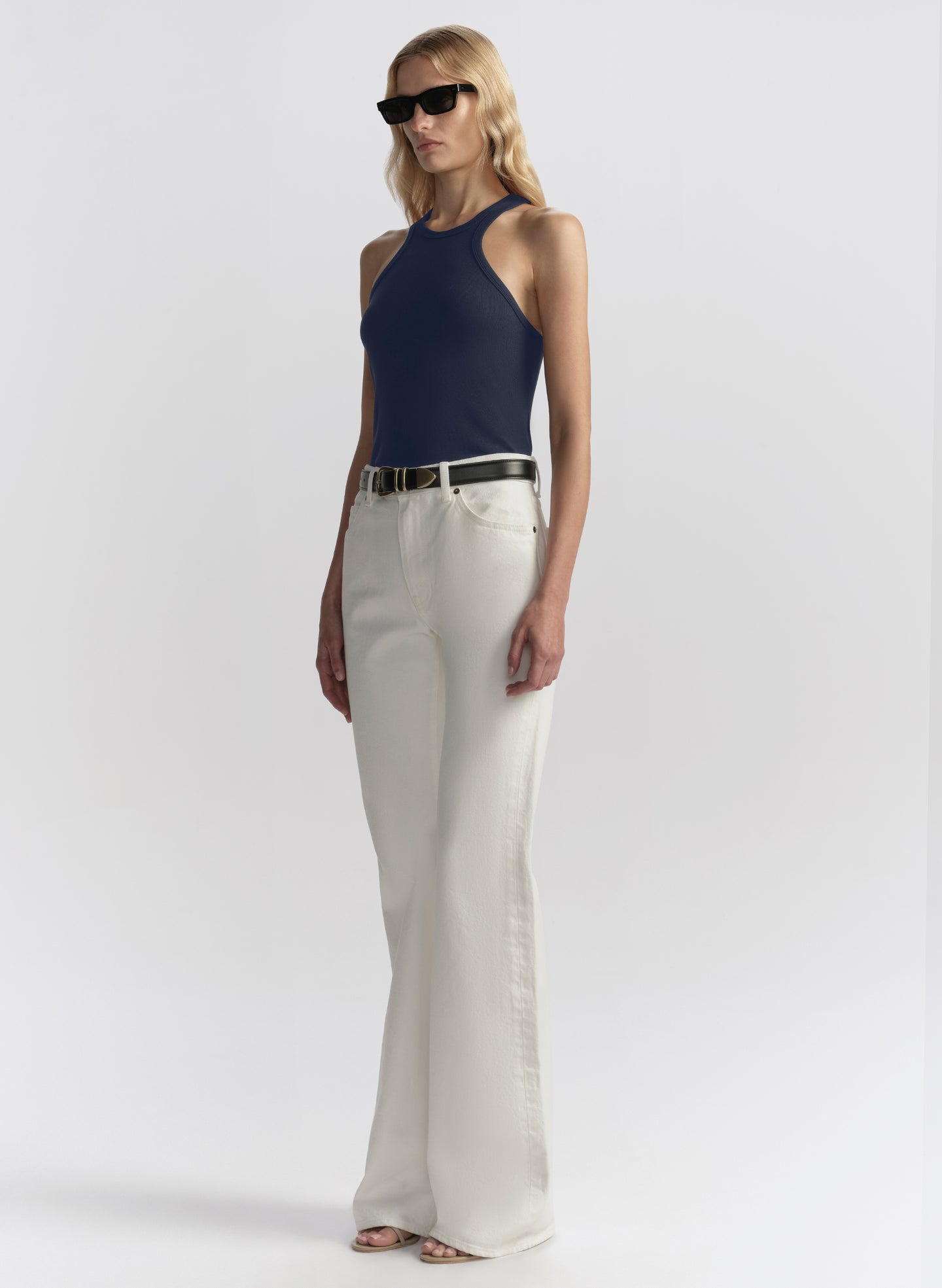 A blonde model wearing a navy blue tank top, off-white wide-leg trousers, a black belt, and black sunglasses, standing against a light gray studio background.