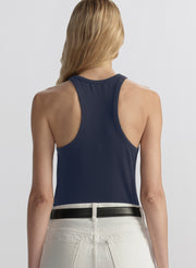 A woman viewed from the back wearing a navy blue racerback tank top and white pants with a black belt, against a light gray studio background.