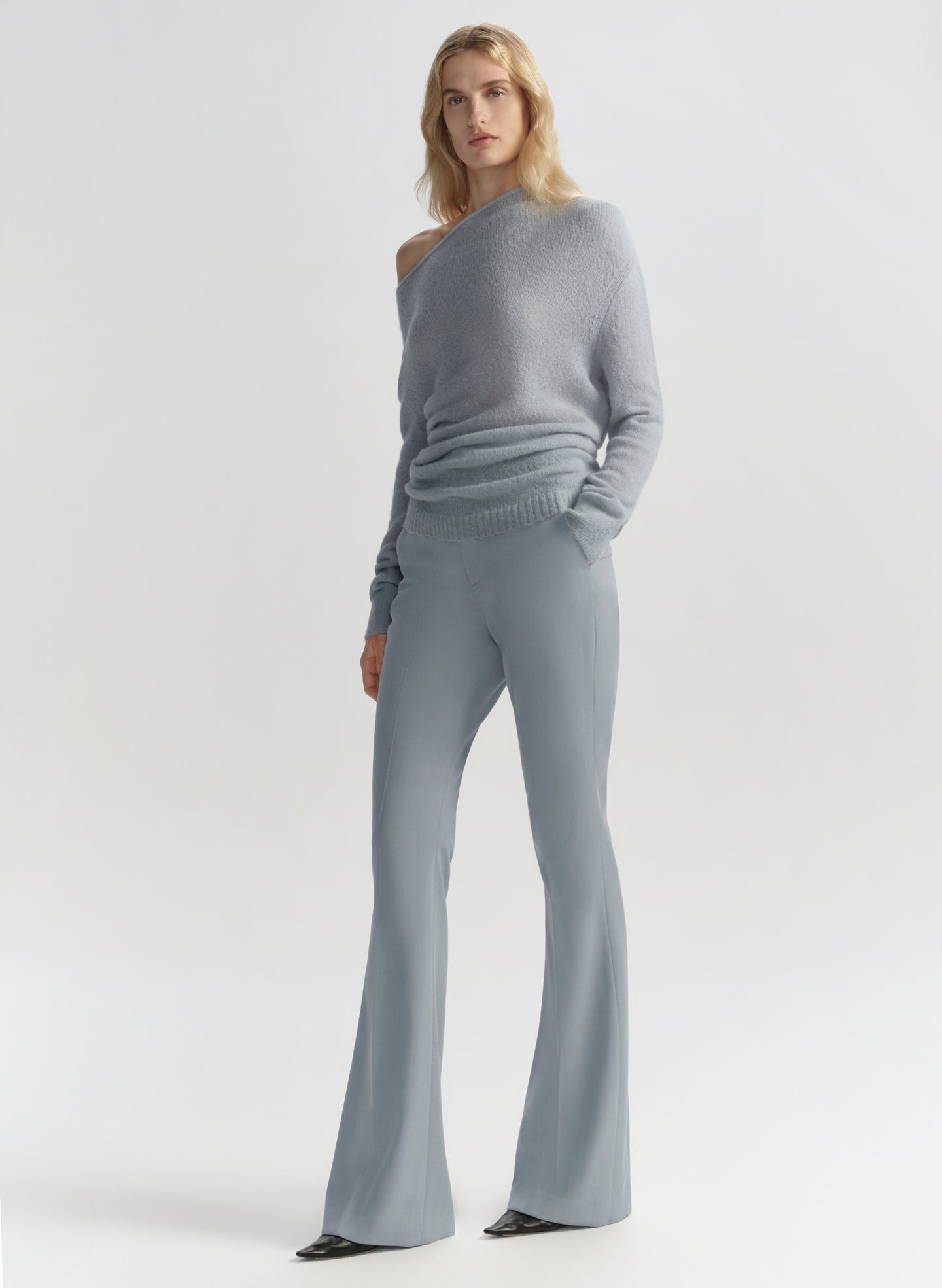 Woman wearing a gray one-shoulder sweater and flared pants on a light gray background
