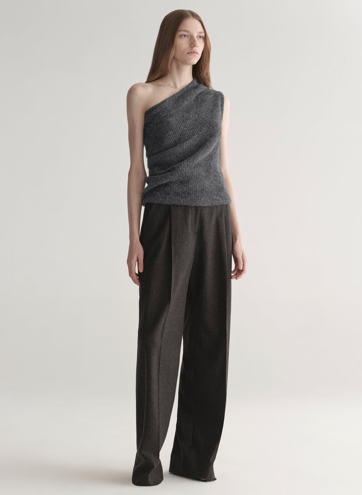 Woman wearing a gray one-shoulder top and dark grey pants on a plain background