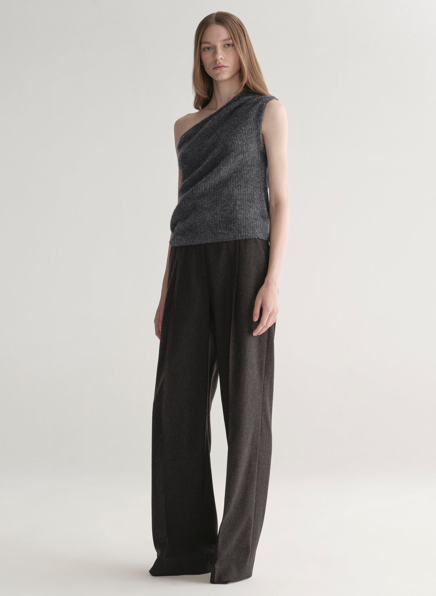 Woman wearing a gray one-shoulder top and dark grey pants on a plain background