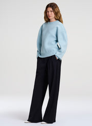 A woman modeling a light blue crewneck sweater and navy wide-leg trousers against a plain light gray studio background.