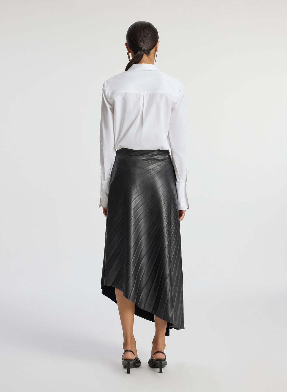 back view of woman wearing white button down top and black pleated vegan leather skirt
